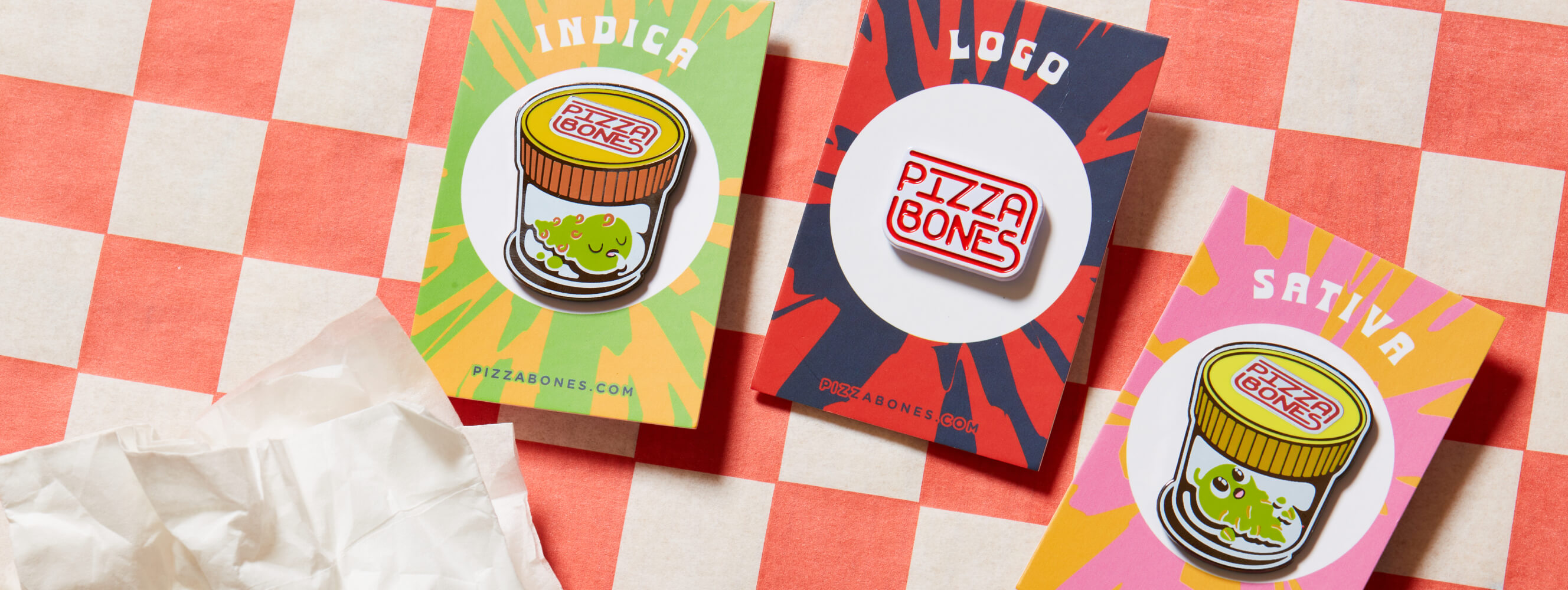 Photo of three Pizza Bones pins, Indica, Pizza Bones Logo, and Sativa, sitting on a backing card. The background is checkered red and white, and there's a crumpled napkin nearby.